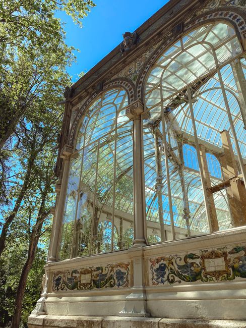 Close-up view of a large, historic greenhouse with ornate metal framing and multiple glass panels, situated in a park setting. The structure features arched windows and detailed decorative elements al