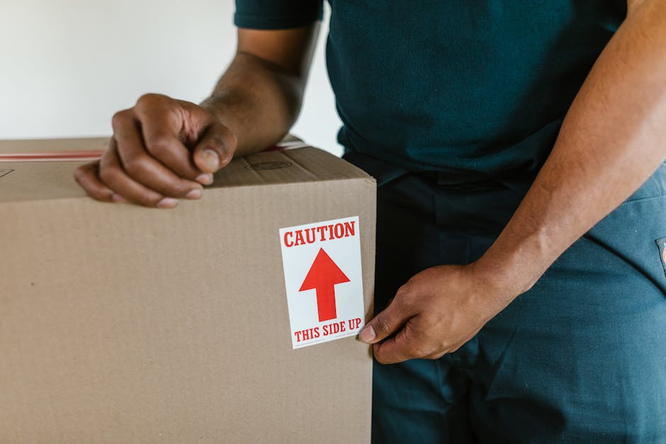 Close-up of a person's hands during home relocation, holding a brown cardboard box with a red and white caution label that reads 'CAUTION THIS SIDE UP' and an upward-pointing arrow. The person is wear